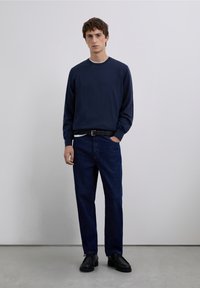 Marinblå tröja med rund halsringning, kombinerad med mörkblå denimbyxor. Svart bälte och skor. Mjuk textur och avslappnad passform. Minimalistisk design.