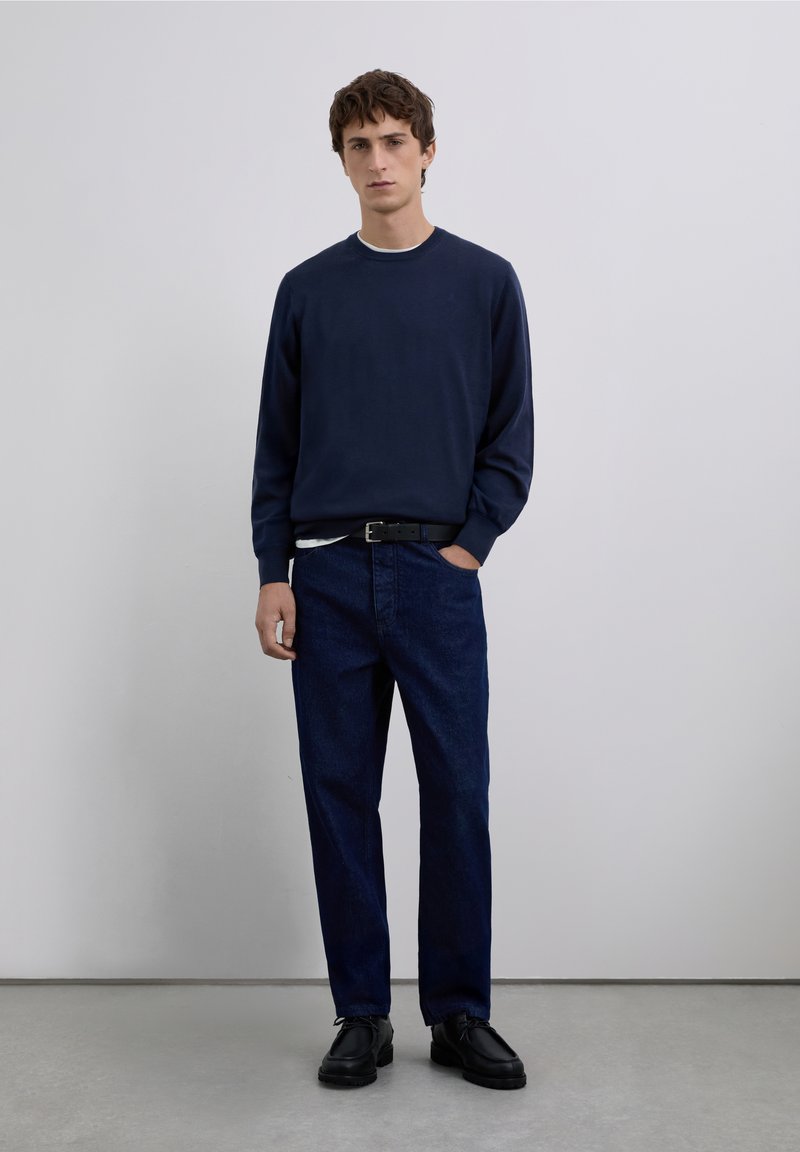 Marinblå tröja med rund halsringning, kombinerad med mörkblå denimbyxor. Svart bälte och skor. Mjuk textur och avslappnad passform. Minimalistisk design.