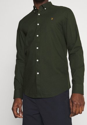 Homme portant une chemise à manches longues boutonnée vert foncé avec un petit "F" jaune brodé sur la poitrine et un pantalon foncé.