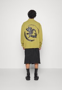 Felpa verde oliva con una grande grafica nera di una creatura mitologica sul retro. Il modello indossa una gonna nera al ginocchio e stivali.