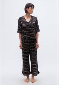 Femme debout, pieds nus, portant une tenue noire deux pièces à pois blancs, avec un grand col et un pantalon large à volants.