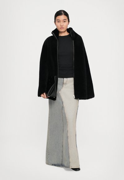 O femeie îmbrăcată cu o jachetă neagră pufoasă cu fermoar, o fustă lungă din denim bicolor, un top negru, ținând în mână o poșetă lucioasă neagră, stând pe un fundal alb.