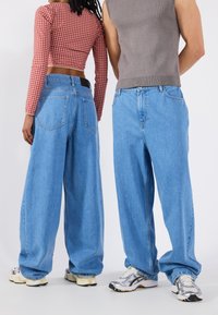 Os jeans de perna larga azuis claros apresentam um design clássico de cinco bolsos. Os indivíduos usam um top curto xadrez vermelho e um top cinza sem mangas.