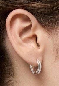 Pendiente de aro de plata con un diseño retorcido, que presenta una textura suave y pulida y una forma circular, exhibido junto a una oreja humana.