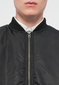 Chaqueta bomber negra con cuello acanalado, cremallera frontal y textura de tela suave. Presenta una camisa blanca con cuello debajo.