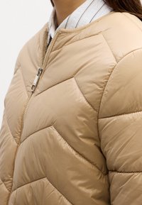 Close-up van een torso met een beige doorgestikte pufferjas met diagonale stiksels en een gedeeltelijk gesloten zilveren rits over een wit overhemd met kraag.