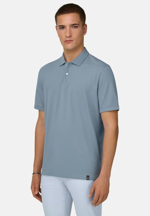 Jonge man met een lichtblauw poloshirt met korte mouwen en een witte broek, staand tegen een effen lichte achtergrond.