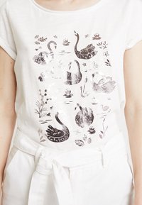 Camiseta de algodón blanca con un estampado de cisne plateado que presenta ocho cisnes, plantas acuáticas y detalles florales. Combinada con pantalones blancos con cinturón.