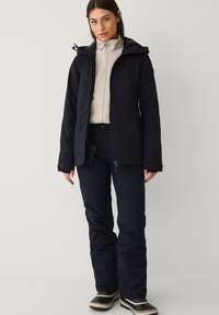 Zwarte waterdichte jas met een capuchon, gecombineerd met een lichtgekleurde fleece met rits en zwarte broek, afgewerkt met zwart-witte laarzen.