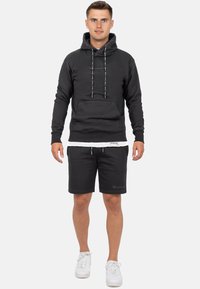 Zwarte fleece hoodie met een borstzak, trekkoorden en logo op de borst, gecombineerd met zwarte shorts en witte sneakers.