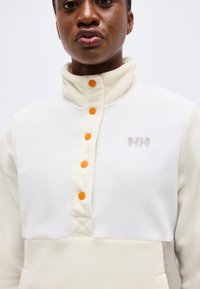 Gros plan d'une personne portant un pull polaire blanc avec des boutons-pression orange et un logo "HH" gris sur la poitrine.