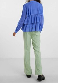 Blusa plissettata blu a strati con maniche lunghe e ruches sulla schiena, abbinata a pantaloni dritti verde chiaro e scarpe con zeppa nere.