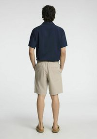 Homem de pé com as mãos nos bolsos, vestindo uma camisa de manga curta azul-marinho, calções bege e sandálias castanhas, de costas para um fundo branco.
