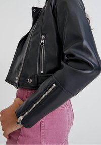 Chaqueta de cuero negra corta con cremalleras plateadas, cierre de botón y bolsillos laterales. Presenta una textura suave y un corte ajustado. Pantalones de mezclilla rosa visibles.
