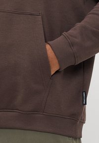 La mano de una persona en el bolsillo delantero de un sudadera marrón con puños acanalados y una etiqueta de marca "PRJCT CORE" en el lado.