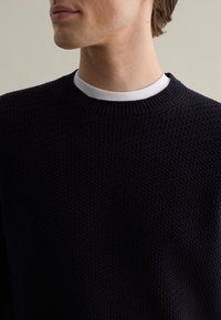 Maglione a maglia navy con un motivo a waffle testurizzato. Presenta un collo rotondo e sovrapposto a una maglietta bianca visibile al colletto.