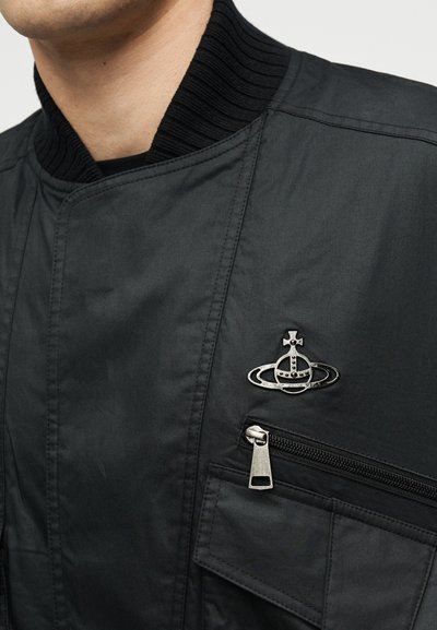 Μαύρο μπουφάν με ριμπ κολάρο, τσέπη στο στήθος με φερμουάρ, τσέπη με καπάκι και ασημένιο έμβλημα με έναν σφαίρα με στέμμα και δακτυλίους στο αριστερό στήθος.