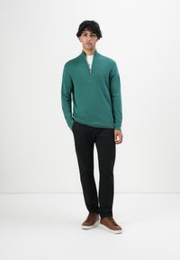 Junger Mann steht da und trägt einen türkisfarbenen Zip-Up-Pullover, schwarze Hosen, ein weißes Unterhemd und braune Sneakers vor einem schlichten weißen Hintergrund.