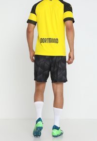 Gul fotbollströja med svarta accenter och "DORTMUND" tryckt på baksidan, kombinerad med svart mönstrade shorts och gröna fotbollsskor.