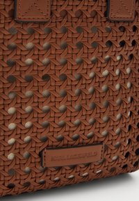 Brązowa pleciona torba o teksturowanym wzorze, z skórzaną metką. Wzór składa się ze skomplikowanych splotów krzyżowych, co nadaje jej szczegółowy wygląd.