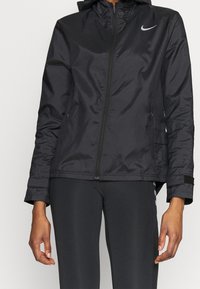 Veste coupe-vent noire avec capuche, dotée d'une fermeture éclair, d'un tissu texturé et d'un logo Nike réfléchissant sur le haut à gauche de la poitrine.