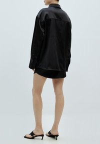 Schwarzes satinoversized Hemd mit Kragen, Knöpfen an den Manschetten und abgerundetem Saum, kombiniert mit einem schwarzen Minirock und Sandalen mit Absatz.