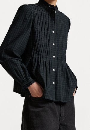 Blusa a maniche lunghe a quadri scuri con dettagli plissettati, chiusura frontale con bottoni, colletto e polsini rifiniti in pizzo, indossata con pantaloni neri e mani nelle tasche.
