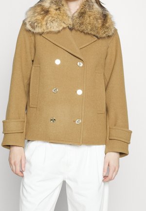 Frau trägt einen zweireihigen Mantel in Kamelbraun mit Kunstpelzkragen und goldenen Knöpfen, kombiniert mit weiten weißen Hosen.