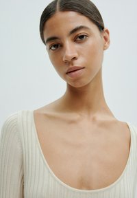 Geribbelde, off-white top met een wijde halslijn, een iets getailleerd ontwerp en lange mouwen. Gladde textuur met een subtiele glans.