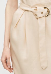 Μπεζ φούστα από ύφασμα με πιέτες μπροστά και ταιριαστή ζώνη με στρογγυλή χρυσή αγκράφα, δίπλα σε ένα χαλαρό χέρι στο πλάι.