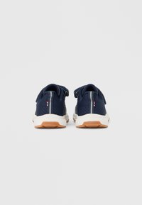 Ζευγάρι αθλητικά παπούτσια σε ναυτικό μπλε χρώμα με λευκές ενδιάμεσες σόλες και εξωτερικές σόλες χρώματος καραμέλας, που προβάλλονται από πίσω πάνω σε λευκό φόντο.