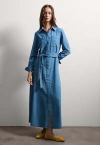 Abito lungo in denim azzurro chiaro, con bottoni frontali, due tasche sul petto e vita legata. Presenta maniche lunghe e una spaccatura laterale.