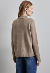 Pull beige avec un col et des poignets côtelés, fabriqué en matériau doux, présentant une coupe décontractée et un ourlet légèrement arrondi, associé à un jean bleu.