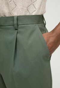 Roheline kohandatud pükste komplekt, mis on valmistatud siledast kangast, pleeditud disainiga, küljetaskudega ja sobiva vöökohtaga. Üleval on nähtav tekstureeritud hele top.