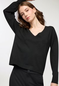 Młoda kobieta z falującymi, brązowymi włosami, nosząca czarny sweter w serek i czarne spodnie, pozująca z uniesioną jedną ręką za głową.