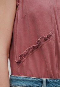 Μπλούζα από καμπαρντίνα σε σκόπινο ροζ χρώμα χωρίς μανίκια, με διαγώνια λεπτομέρεια βολάν κοντά στη μέση, σε συνδυασμό με μπλε τζιν παντελόνι.