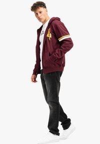 Bordová mikina na zips s rukávmi v žltých a bielych pruhoch, predné vrecká, v kombinácii s čiernymi džínsami a bielymi teniskami.