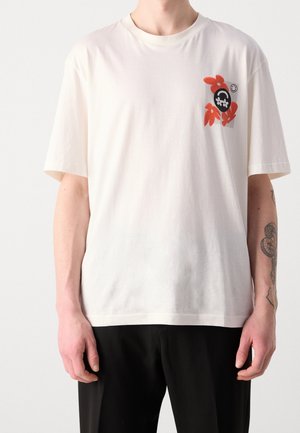 Personne portant un T-shirt blanc avec un petit graphique abstrait rouge et noir ainsi qu'un visage souriant sur le côté gauche de la poitrine, associé à un pantalon noir.
