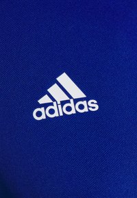 Logo blanc Adidas avec trois bandes diagonales sur un fond en tissu bleu texturé.