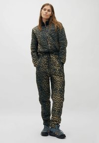 Frau steht mit Händen in den Taschen, trägt eine Jacke mit Leopardenmuster und passende Hose sowie Wanderschuhe vor einem schlichten Hintergrund.
