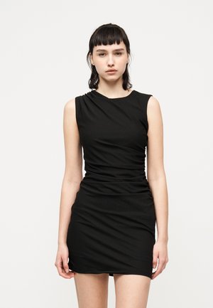 Młoda kobieta o ciemnych włosach, ubrana w bezrękawową czarną marszczoną mini sukienkę, stojąca na tle jednolitego białego tła.