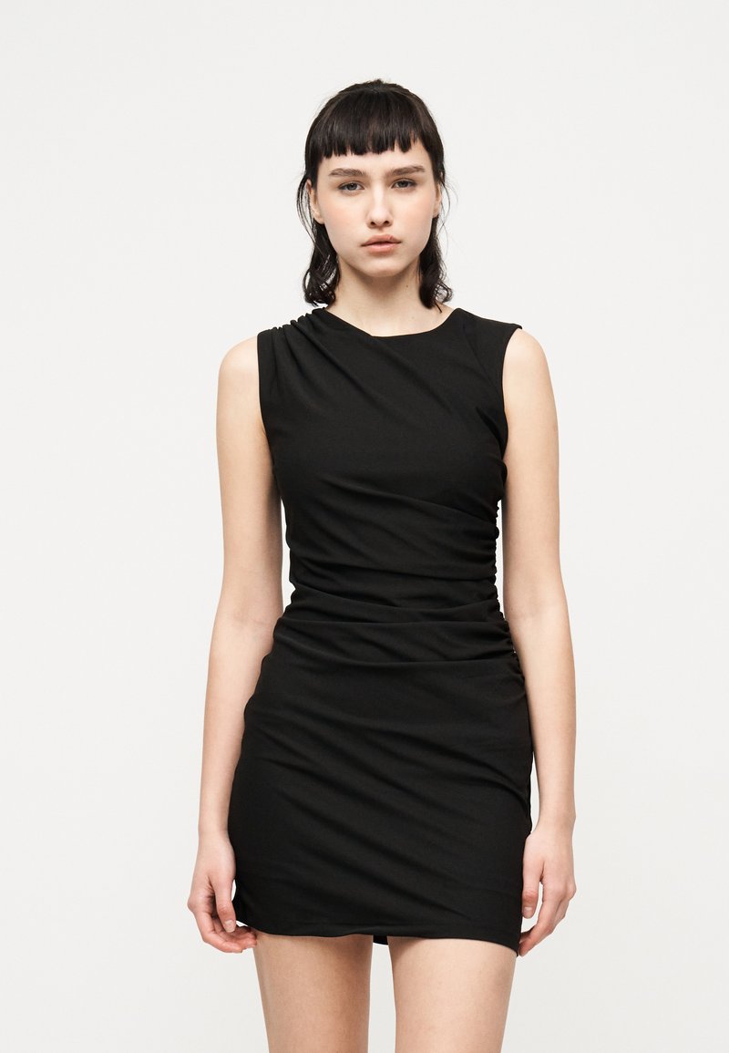 Νεαρή γυναίκα με σκούρα μαλλιά, ντυμένη με μια μίνι μαύρη ριγέ αμάνικη φόρεμα, στέκεται μπροστά σε λευκό απλό φόντο.