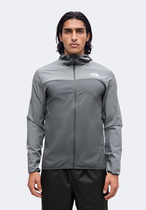 Mand iført en grå lynlåsjakke med hætte og The North Face-logo på brystet, stående mod en ensfarvet baggrund.