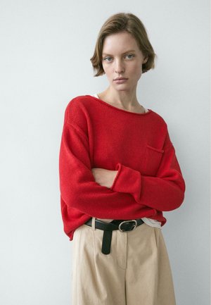 Giovane con capelli corti che indossa un maglione rosso e pantaloni beige, con le braccia incrociate, in piedi davanti a uno sfondo chiaro e uniforme.