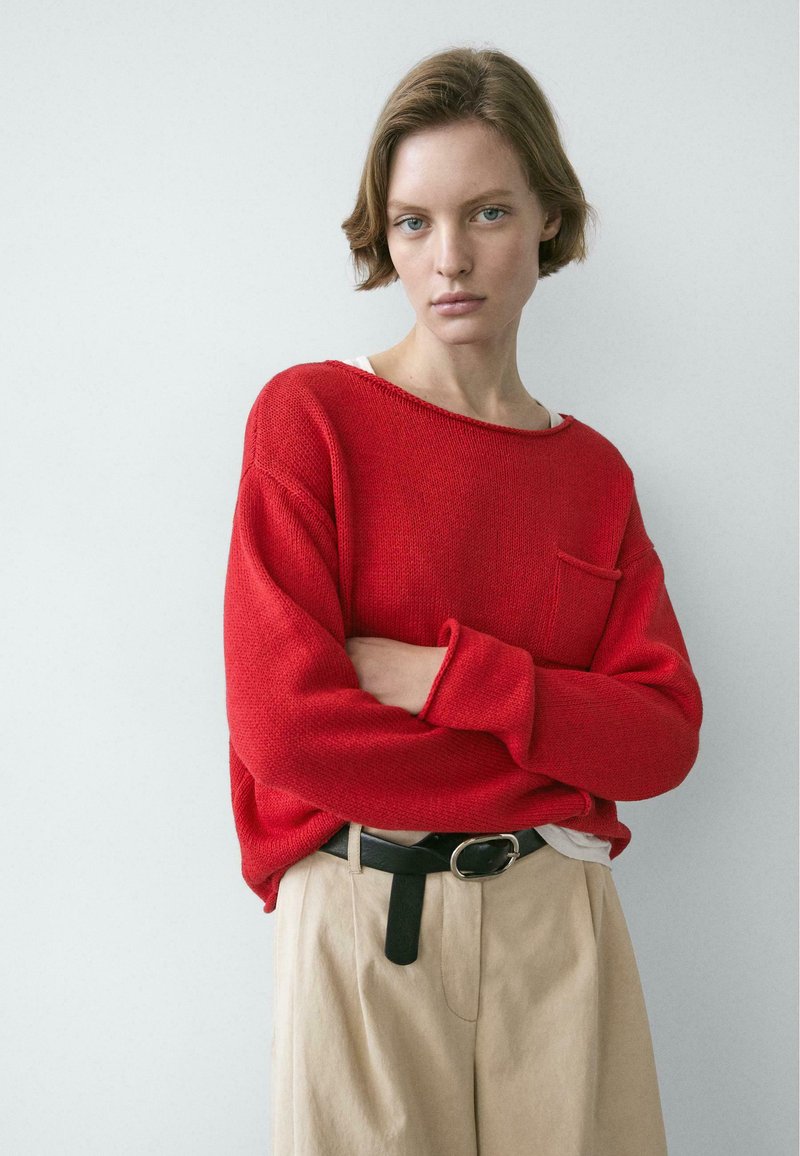 Giovane con capelli corti che indossa un maglione rosso e pantaloni beige, con le braccia incrociate, in piedi davanti a uno sfondo chiaro e uniforme.