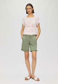 T-shirt rose clair à manches courtes avec un texte graphique rouge et des formes, associée à un short vert retroussé et des sandales blanches avec un accent décoratif.