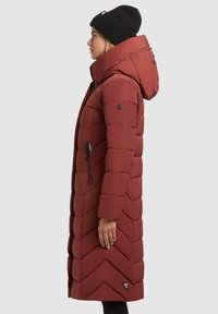 Vrouw in een lange roestkleurige gewatteerde jas met een capuchon en een zwarte baret, staand in profiel tegen een effen achtergrond.