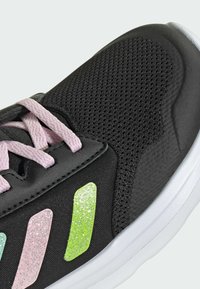 Chaussure de sport noire avec une texture en maille, lacets roses et trois accents scintillants en vert, rose et bleu clair sur le côté. Semelle blanche.