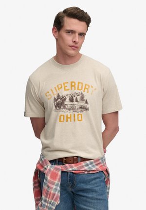 Ung mand iført en beige Superdry Ohio t-shirt, blå jeans og en rød ternet skjorte bundet om livet, stående mod en ensfarvet baggrund.