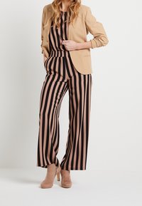 Blazer beige sobre un mono a rayas negro y rosa con cintura elástica; modelo con zapatos de tacón nude; textura del tejido suave.